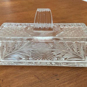 Peut inclure: Beurrier en verre transparent avec une base et un couvercle rectangulaires. Le couvercle est doté d'une poignée et de côtés en forme de losange. La base et le couvercle sont décorés d'un motif floral.