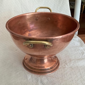 May include: A large, round copper bowl with a polished finish, featuring a wide, flared rim and a sturdy pedestal base. Two curved brass handles are securely attached to the sides of the bowl with visible rivets, providing a functional grip.