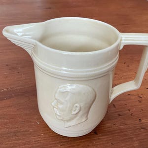 May include: Cream-coloured ceramic jug with a spout and a rectangular handle. The jug features a raised relief profile of a man's face on the front. The jug has a classic, vintage design.