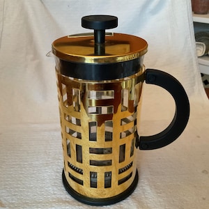 May include: A gold-coloured French press coffee maker with a black handle and lid. The metal cage has a geometric pattern. The glass carafe is visible through the gold cage. The lid has a black knob.