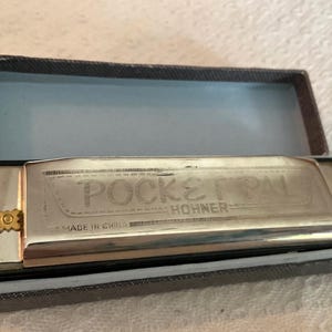 May include: A silver-toned Hohner Pocket Pal harmonica with gold-coloured side screws, resting inside a rectangular dark grey case. The metal surface of the harmonica is engraved with the text POCKET PAL, HOHNER, and MADE IN CHINA.