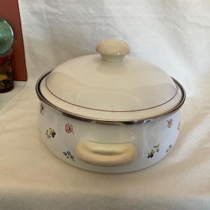 Peut inclure: Une casserole en émail blanc avec un motif floral et un couvercle de couleur crème. La casserole a une poignée de couleur crème.
