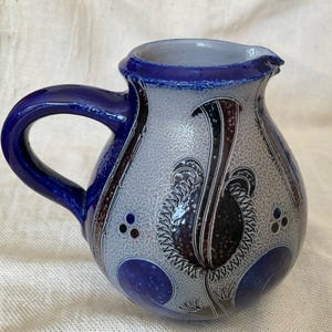 Peut inclure: Une cruche en céramique avec une anse et un bord bleus. Le corps est gris clair avec un motif texturé et des motifs floraux bleu foncé, noirs et marron. La cruche a un bec verseur.