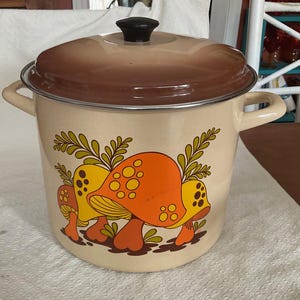May include: Cream enamel pot with a brown lid and black handle. The pot has a whimsical design of orange and yellow mushrooms with green foliage. A vintage kitchenware item.