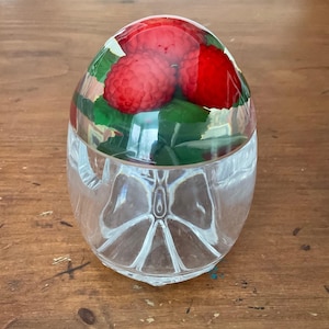 Peut inclure: Un presse-papier en verre transparent avec un dessus arrondi, contenant trois framboises rouges vives et des feuilles vertes. La base du presse-papier présente un motif décoratif de papillon.