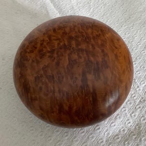 以下が含まれることがあります： 滑らかで光沢のある仕上げの円形の木製ボックス。蓋は、濃い茶色の複雑で渦巻くようなバールウッドの木目模様が特徴で、表面全体に暗く複雑な節と有機的な大理石のような質感が広がっています。