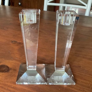 Peut inclure: Deux bougeoirs géométriques en cristal transparent. Chaque bougeoir a une base carrée, une tige effilée et une coupelle carrée pour une bougie. Les bougeoirs sont en cristal transparent et sont présentés sur une surface en bois.