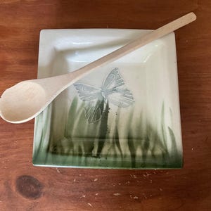 Peut inclure: Un repose-cuillère carré en céramique avec un motif de papillon et une cuillère en bois. Le repose-cuillère est blanc avec de l'herbe verte et un papillon gris. La cuillère en bois est brun clair.