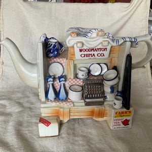May include: A white ceramic teapot shaped like a shop front with the words "Woodmanton China Co." on the front. The teapot has a blue and white striped awning and a sign that says "Carr's Finest Tea Pots".