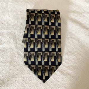 Peut inclure: Une cravate pliée avec un motif géométrique répétitif. La cravate présente un fond bleu marine foncé avec un motif répétitif de formes beiges, bleu clair et noires. La cravate est en tissu lisse.