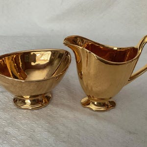 Peut inclure: Un ensemble de crémier et sucrier dorés. Le pot à crème a une poignée incurvée et un bec verseur. Le sucrier est de forme ovale. Les deux pièces ont une surface texturée et reposent sur une base décorative. L'ensemble est en matériau brillant et réfléchissant.