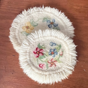 May include: Two cream-coloured, round coasters with fringed edges. Each coaster features a floral design in shades of pink, blue, and yellow, with green leaves. The coasters are made of a textured, woven material.