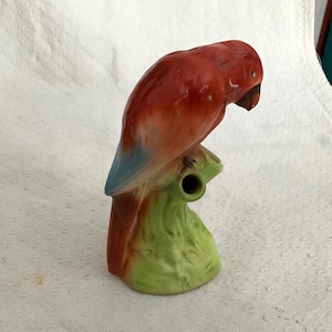 May include: A ceramic parrot figurine. The parrot has a red body, blue wing accents, and a brown beak. It is perched on a green branch. The figurine is likely a decorative item.