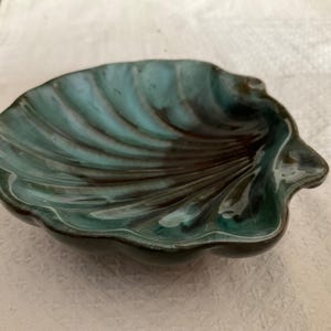 May include: A ceramic dish shaped like a seashell with a glossy finish. The interior features a blend of deep teal and dark brown glaze, with raised ridges radiating from the centre to the scalloped edges, creating a textured, shell-like appearance.