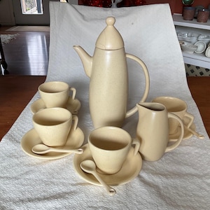 May include: A cream-coloured coffee set with a speckled finish. The set includes a tall coffee pot with a lid, several cups, saucers, spoons, and a creamer. The set is arranged on a white tablecloth.