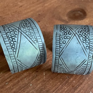 May include: Two silver metal cuff bracelets with detailed geometric patterns. The designs include circles, lines, and diamond shapes. The bracelets are displayed on a wooden surface.
