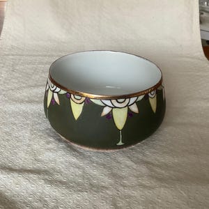 May include: A ceramic bowl with a dark green exterior and a white interior. The rim is accented with gold and a repeating pattern of yellow, white, and purple floral designs. The bowl has a unique shape and is suitable for serving or display.