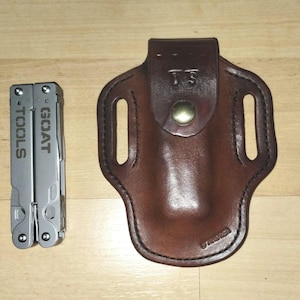 May include: A brown leather sheath with a belt loop and a snap closure. The sheath is designed to hold a multi-tool. The multi-tool is silver and has the words "GOAT TOOLS" engraved on it.
