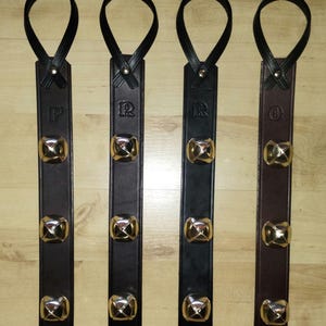 Könnte beinhalten: Vier Lederriemen mit goldfarbenen Glocken und schwarzen Schlaufen. Die Riemen sind in Braun- und Schwarztönen gehalten und mit den Buchstaben "F", "R", "R" und "O" geprägt. Die Glocken sind quadratisch und haben ein glänzendes Finish.