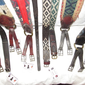 May include: A collection of eight leather cinch straps with different colored woven patterns. The straps have brass buckles and are made by "Buckaroo Leather".