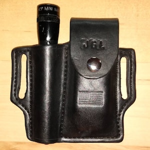 Könnte beinhalten: Ein schwarzes Lederholster mit Druckknopfverschluss und einem amerikanischen Flaggenmuster. Das Holster ist für eine kleine Taschenlampe ausgelegt. Die Taschenlampe ist schwarz und trägt den Text "MINI MAG" auf sich.