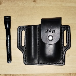 May include: Black leather holster with two slots for carrying a torch and a knife. The holster has a belt loop and the text "KCM" is embossed on the front.