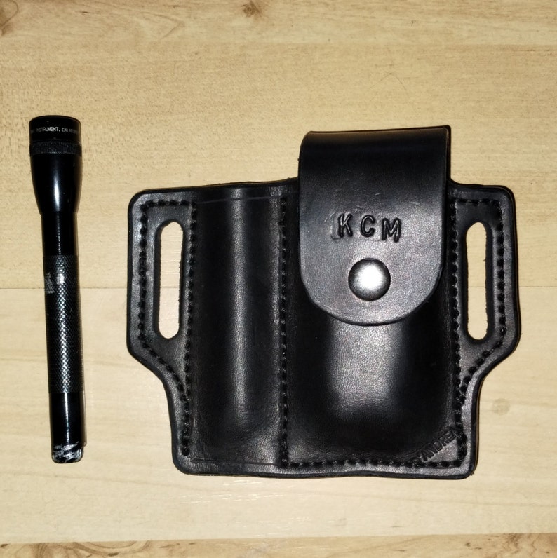 May include: Black leather holster with two slots for carrying a torch and a knife. The holster has a belt loop and the text "KCM" is embossed on the front.