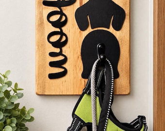May include: A rectangular light brown wooden wall plaque featuring the black text "woof" in a cursive font and a black silhouette of a dog head and body. A black metal hook is attached to the center, holding a lime green and black dog harness.