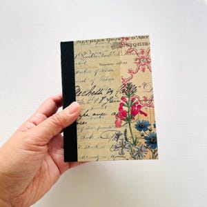 May include: A small notebook with a black spine and a floral design. The cover features a vintage script background with pink and blue flowers. The notebook is held in a hand.