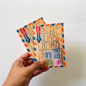 May include: Two vintage-style air mail envelopes featuring a classic red and blue striped border. The envelopes are printed with aged paper textures, black postmarks, handwritten script, and various postage stamps, including the text AIR MAIL.