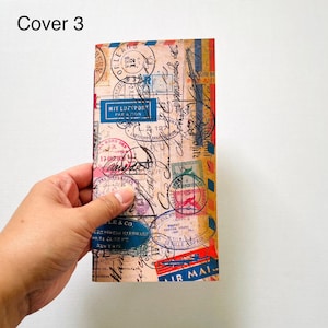 May include: A rectangular notebook cover featuring a vintage airmail theme with a beige background, various black ink postmarks, stamps, and handwritten script. The design includes blue, red, and yellow accents with text such as MIT LUFTPOST.