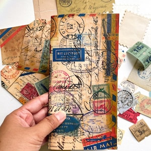 May include: A vintage-style paper journal with a collage of old postage stamps, airmail labels, and handwritten notes. The cover features a blue stamp that reads "MIT LUF POST PAR ANON".