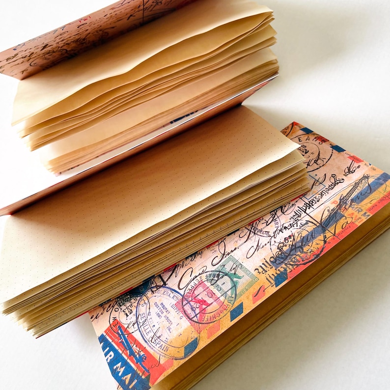 May include: A notebook with a vintage airmail-themed cover featuring postage stamps, postmarks, and handwritten script. The notebook is open, revealing multiple stacks of light tan, dotted paper pages bound together in a layered, accordion-style format.