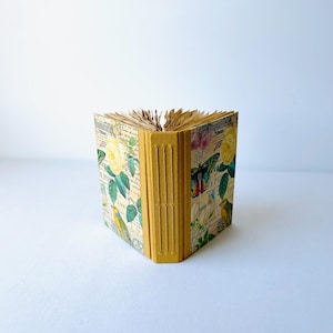 May include: A small, yellow, handmade journal with a floral and bird design on the cover. The journal has a spine with five raised bands.
