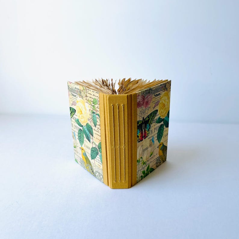 May include: A small, yellow, handmade journal with a floral and bird design on the cover. The journal has a spine with five raised bands.