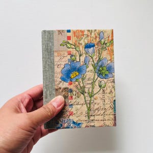 May include: A small, handmade journal with a linen cover and a floral design. The cover features blue flowers with yellow centers and green leaves on a background of vintage-looking paper with handwritten text.