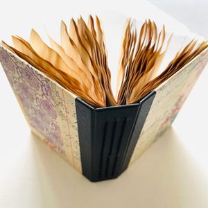 May include: An open journal with aged paper pages. The cover features a vintage floral and script design with purple and pink accents. The spine is black, and the pages are a warm, sepia tone.
