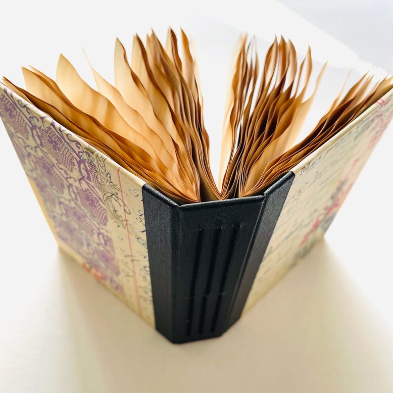 May include: An open journal with aged paper pages. The cover features a vintage floral and script design with purple and pink accents. The spine is black, and the pages are a warm, sepia tone.