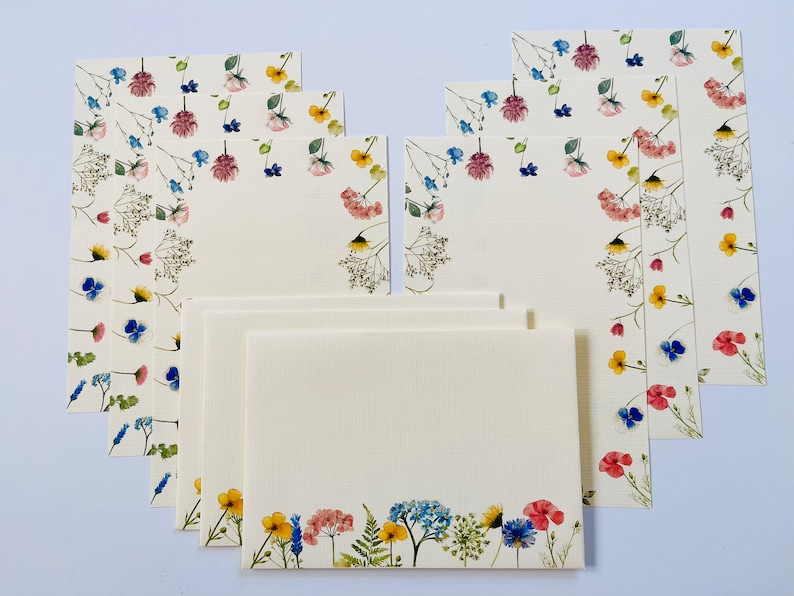 May include: A set of eight blank note cards with a floral border. The cards are white with a delicate pattern of pressed flowers in shades of pink, blue, yellow, and green.