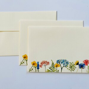 May include: Three blank ivory note cards with a border of pressed flowers in shades of yellow, blue, and pink. The flowers include a yellow buttercup, blue forget-me-nots, and pink Queen Anne's Lace.