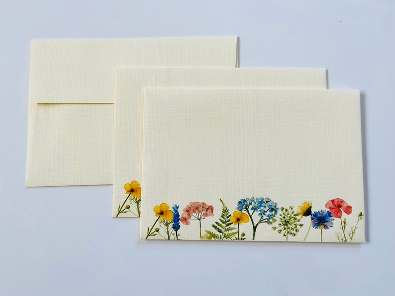 May include: Three blank ivory note cards with a border of pressed flowers in shades of yellow, blue, and pink. The flowers include a yellow buttercup, blue forget-me-nots, and pink Queen Anne's Lace.