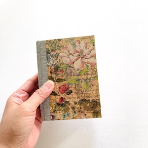 Puede incluir: Un pequeño cuaderno con estampado floral y cubierta de lino gris. La cubierta presenta una gran flor rosa con hojas verdes y un fondo de texto escrito a mano.