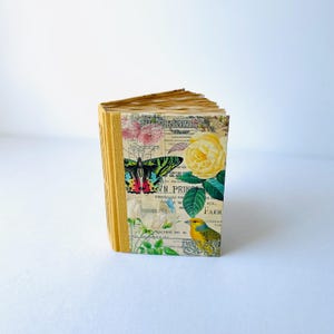 May include: A small, yellow journal with a floral design. The cover features a butterfly, a rose, and a bird. The text on the cover reads "The Perennial Farm".