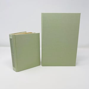 Puede incluir: Dos libros de color verde salvia con cubiertas de tela. El libro más pequeño a la izquierda presenta un lomo cosido expuesto con hilo visible, mientras que el libro más grande a la derecha tiene una cubierta lisa y sólida. Ambos son rectangulares.