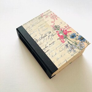 May include: A closed journal with a black spine and pages covered in floral and script designs. The cover displays pink and blue flowers and vintage handwriting. The journal is rectangular and appears new.