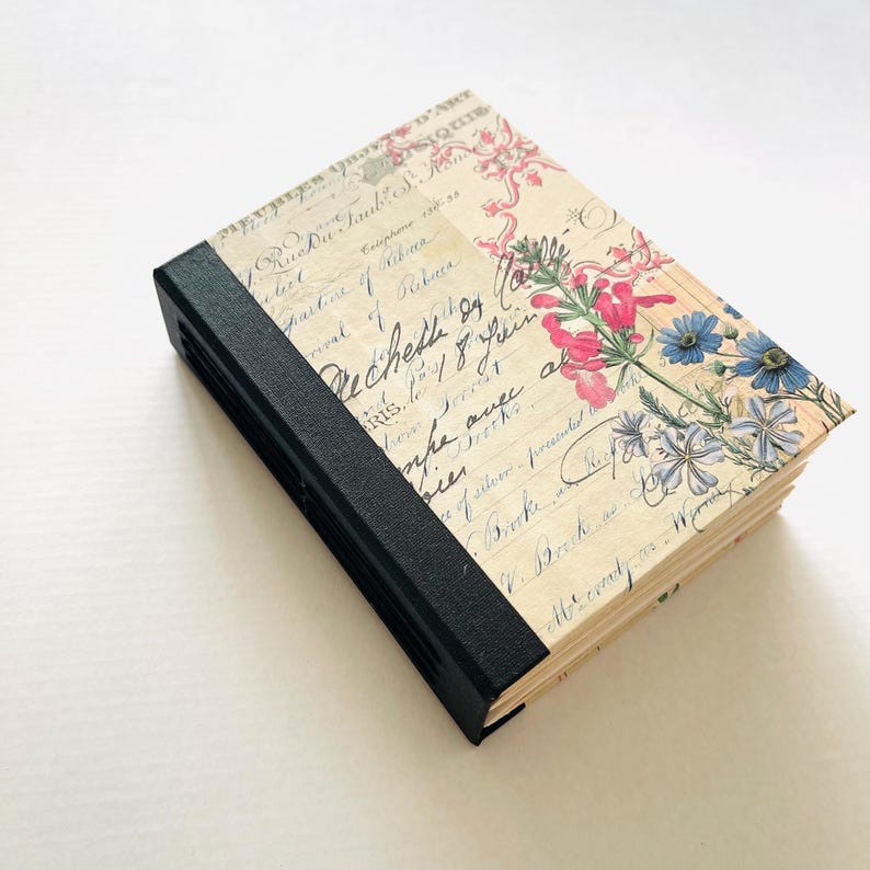 May include: A closed journal with a black spine and pages covered in floral and script designs. The cover displays pink and blue flowers and vintage handwriting. The journal is rectangular and appears new.