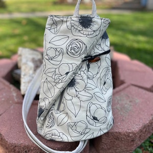 May include: A white canvas backpack with a black floral print. The backpack has a zipper closure and a top handle.