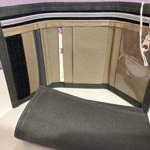 May include: Open, gray trifold wallet with multiple compartments. Features a zippered pocket, clear ID window, and a Velcro closure. The interior is tan and gray, with a white zipper and a silver zipper pull.