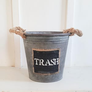 May include: A galvanized metal trash can with rope handles. The can has a burlap patch with the word "TRASH" written in white paint.