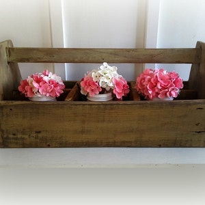 May include: A rustic wooden tool box with a handle, painted in a green and brown distressed finish. Three small white jars are placed inside the box, each filled with pink and white flowers.